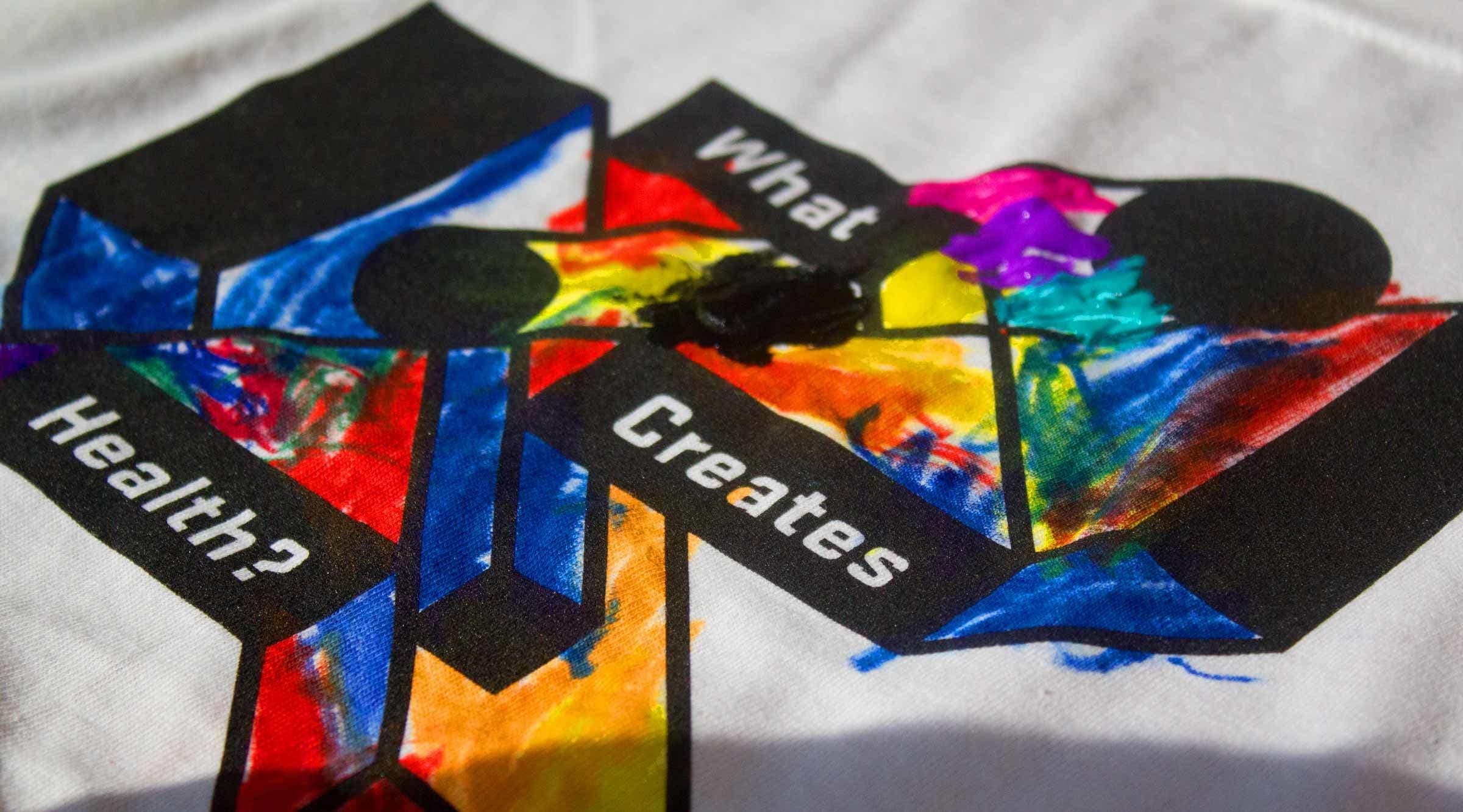 A colorful abstract artwork featuring two black silhouettes facing each other and overlapping. Bright multicolored paint splashes are integrated into and around the shapes. Text on the artwork reads, "What Creates Health?" in bold white letters.