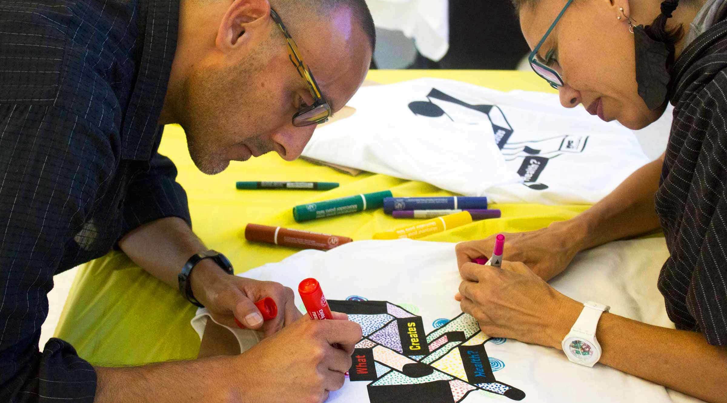 Two people are working together to color a design on a white t-shirt using markers. The design includes geometric shapes and text. Various colored markers are scattered on the yellow tablecloth in front of them. They are focused and wearing glasses.