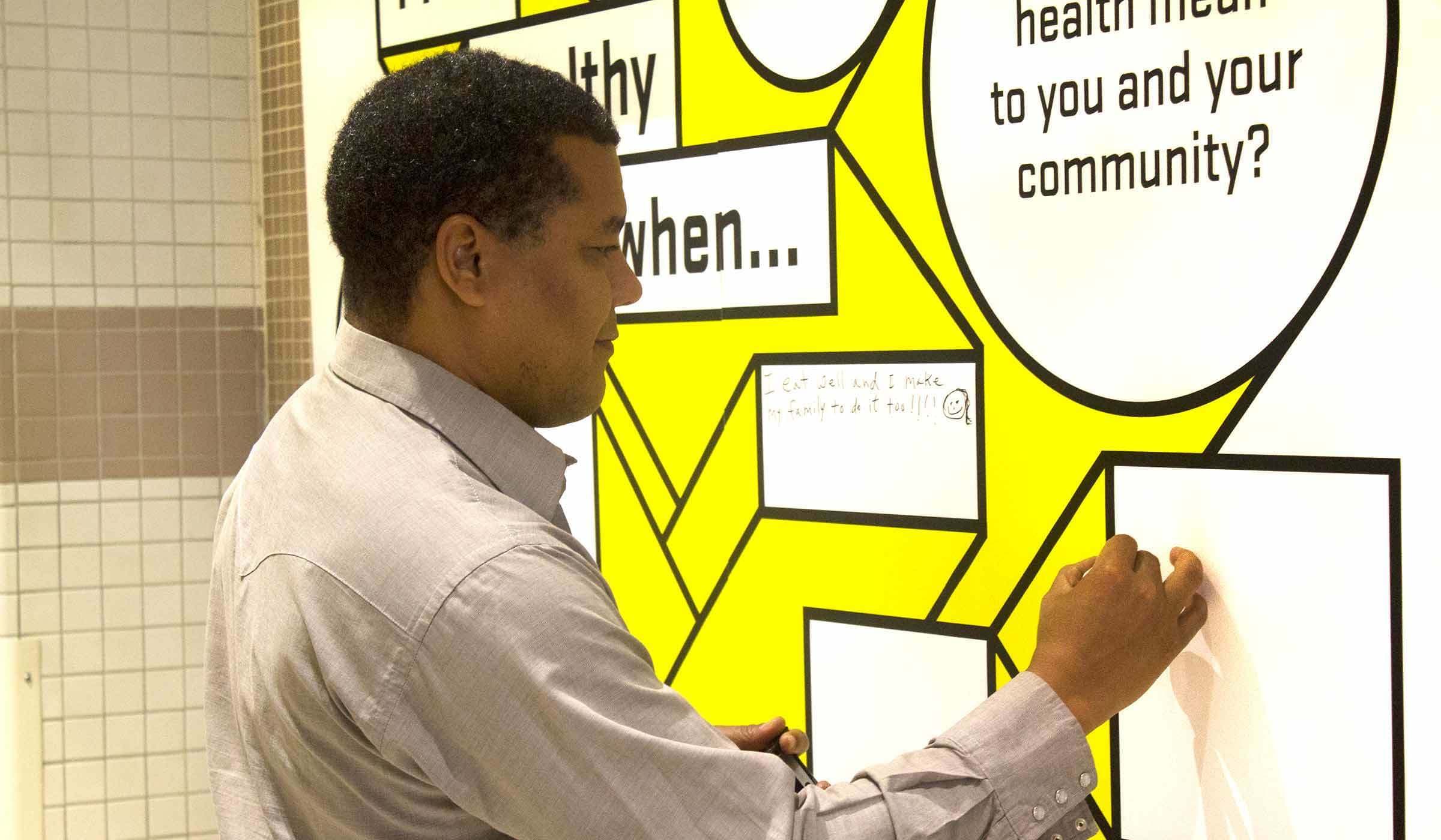 A man in a light gray shirt stands in front of a wall with yellow and white speech bubbles. He is writing on a blank speech bubble with a marker. The background has text, "What does health mean to you and your community?.