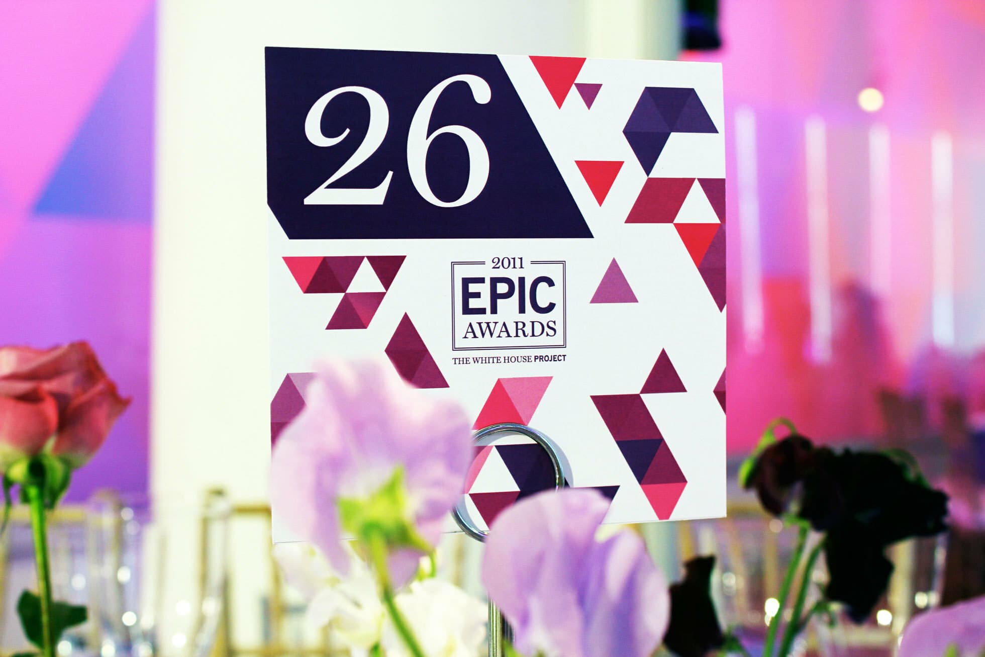 A table number card with "26" and "2011 EPIC Awards The White House Project" printed on it, decorated with a geometric red and purple pattern. The card is placed on a metal holder among flowers, against a pink and purple blurred background.