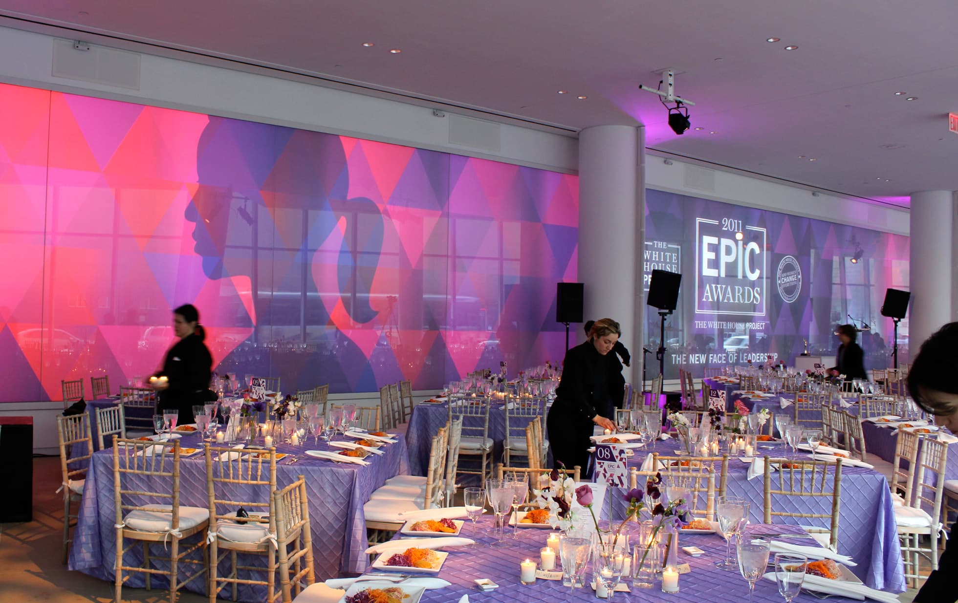 A modern event room with purple and pink lighting features tables set for dining, adorned with candles and floral centerpieces. Two staff members are preparing the setup. "Epic Awards" signage is visible on the large windows in the background.