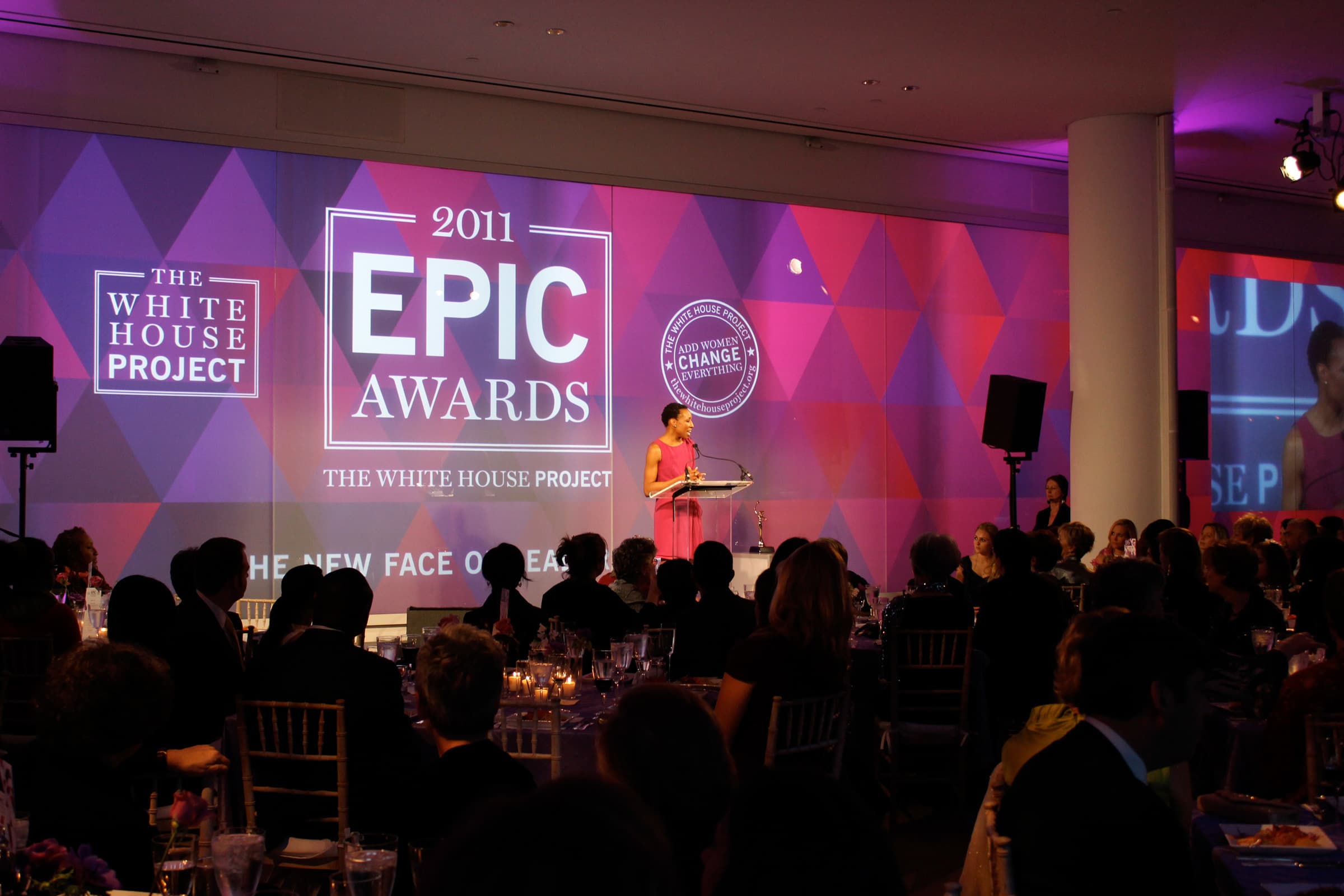 A woman stands at a podium speaking to a seated audience in a formal event setting. On the wall behind her are projected graphics that read "2011 EPIC Awards" and "The White House Project." The stage area is lit with purple and pink lighting.