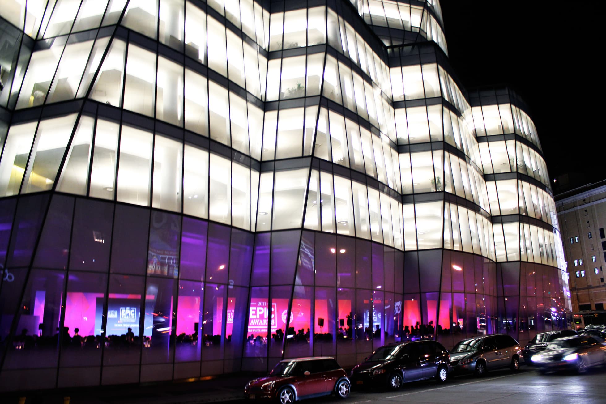 A modern, multi-level building with angular, illuminated glass walls at night. The interior lights create a bright, warm ambience. Several cars are parked along the street in front. The lower level has purple and pink lighting, indicating possible commercial activity.