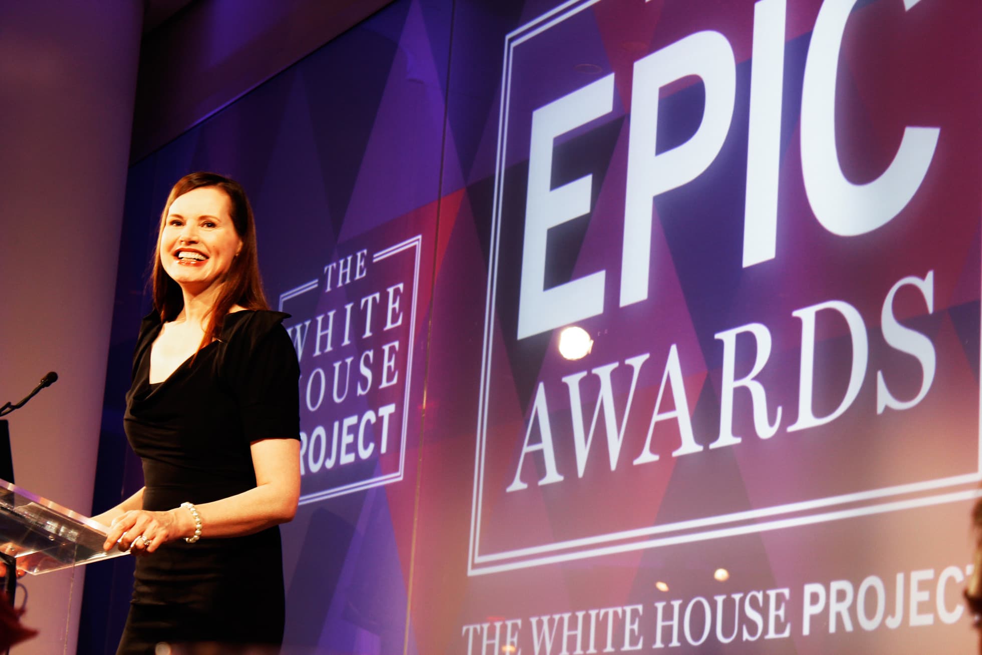A woman in a black dress stands at a podium, smiling. Behind her, signage reads "THE WHITE HOUSE PROJECT" and "EPIC AWARDS." The backdrop features a modern, geometric design with shades of purple and red.