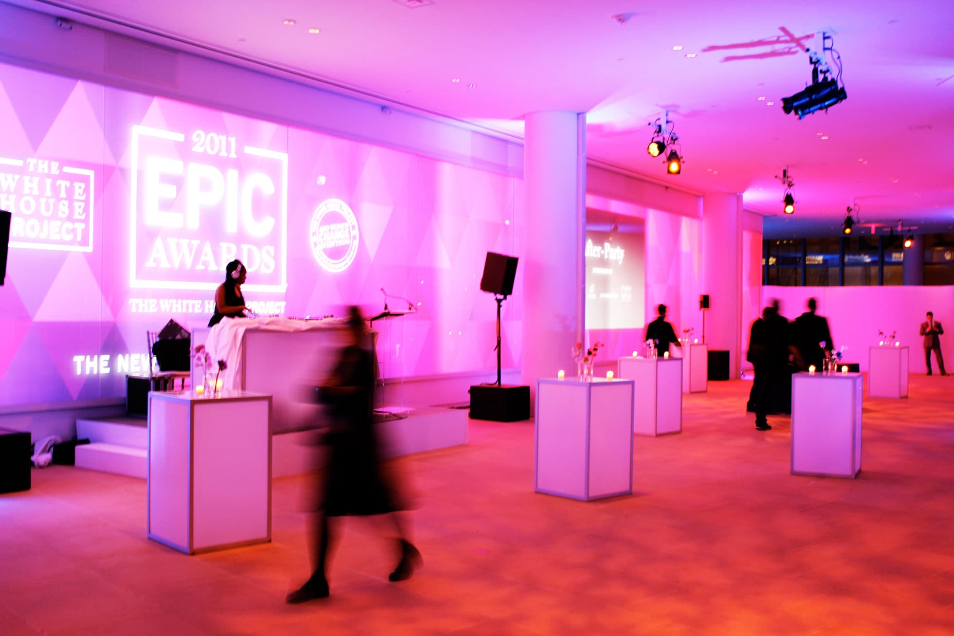 A sleek event hall with purple and pink lighting hosts the 2011 EPIC Awards. High tables with candles are sparsely placed around the room. A person stands at a DJ booth on an elevated platform, while several attendees are blurred in motion.