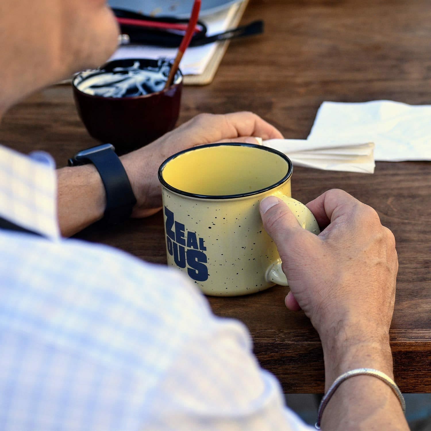 A person with light skin wearing a wristwatch and holding a yellow speckled mug that says "Zeal Us" on it. The person is sitting at a wooden table with a napkin and a red spoon. In the background, there is a bowl filled with sugar packets and some papers.