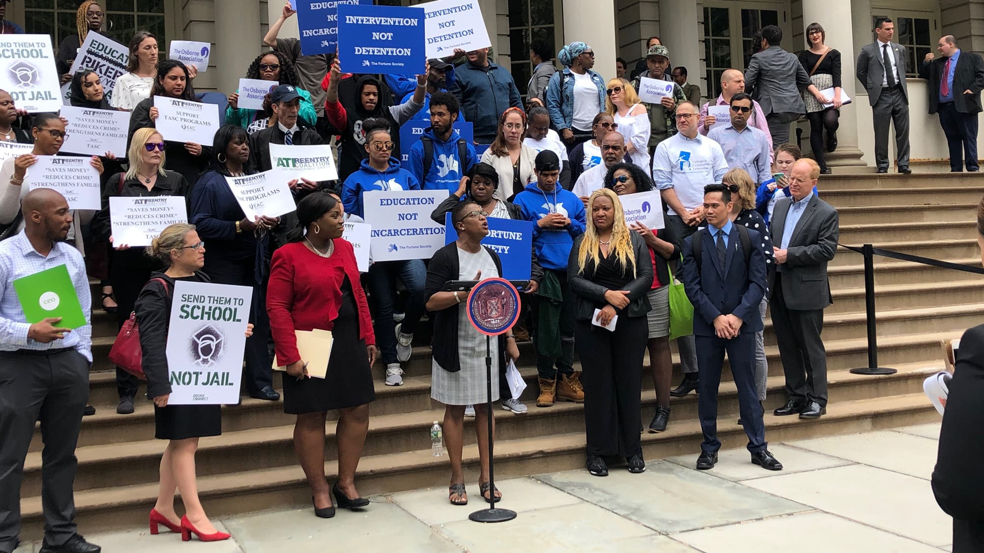 A group of people at a protest on the steps of a building, holding signs with messages like "Education Not Incarceration" and "Send Them to School, Not Jail." A speaker stands behind a podium addressing the crowd, surrounded by supporters and media.