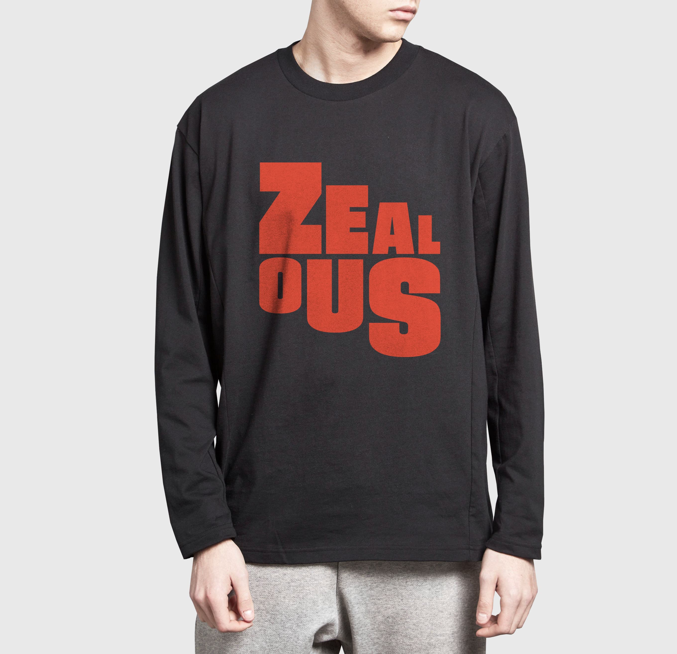 A person wearing a black long-sleeve shirt with the word "ZEALOUS" printed in large red letters on the front stands against a gray background. They are also wearing light gray sweatpants. Only their torso and part of their head are visible.