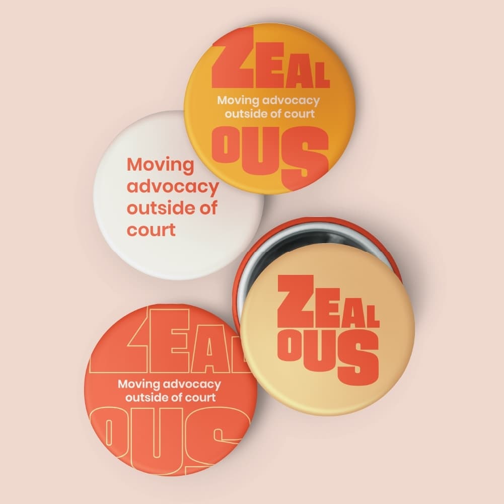 Four circular pins with vibrant designs are displayed on a light pink background. Two pins read "ZEALOUS" in bold letters with "Moving advocacy outside of court" beneath it. Another pin reads "Moving advocacy outside of court" in red text. The fourth pin is partially open, its inscription is obscured. The pins are in yellow, white, and red color schemes.